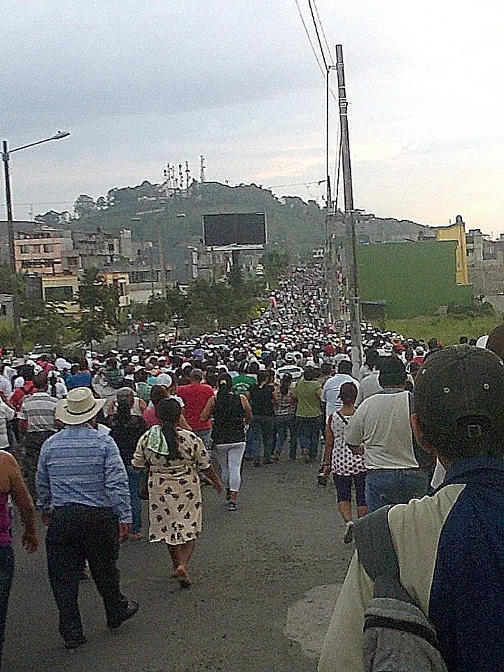 Una multitud de devotos camina por una avenida cuesta arriba, entre postes de luz y casas, en una procesión de la Virgen de El Cisne, con un cerro al fondo