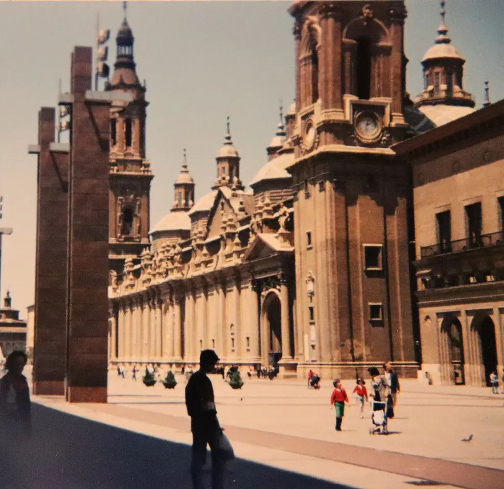 Fachada larga de la basílica del Pilar, de ladrillo, con sus torres y cúpulas, vista desde la plaza soleada, con paseantes y niños en primer plano