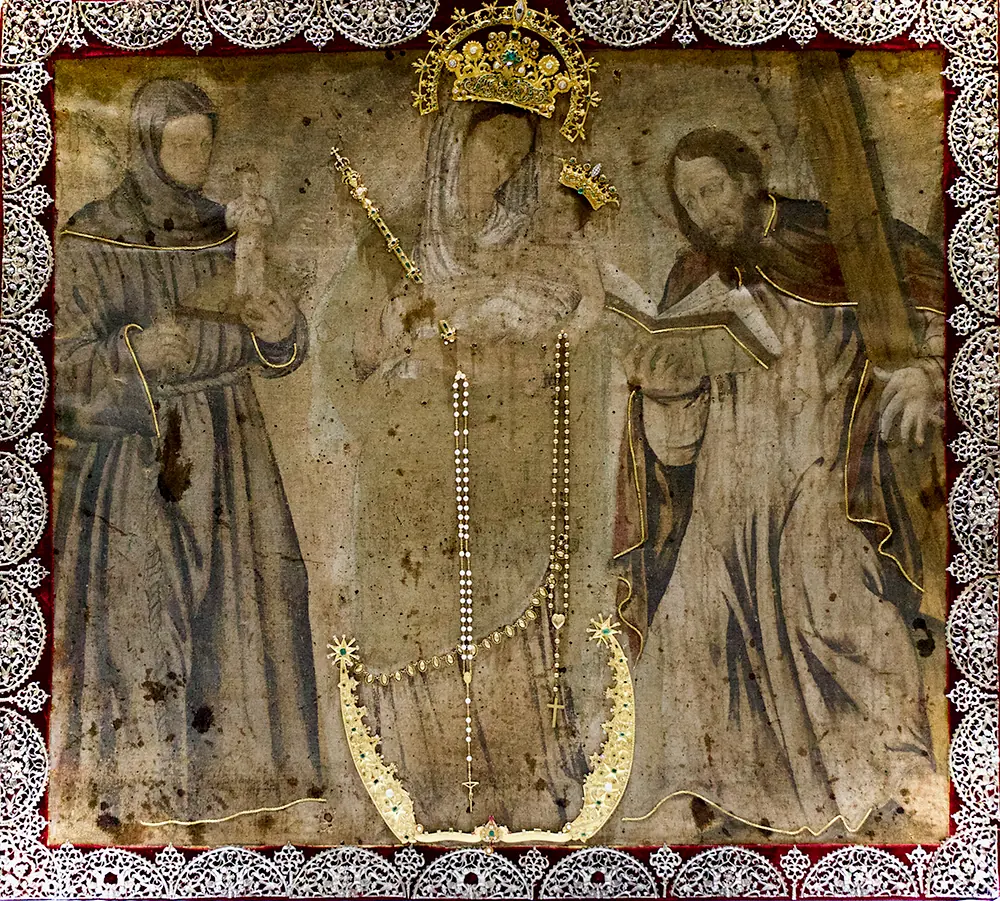 Lienzo antiguo y desgastado en tonos ocre de la Virgen coronada con el Niño y un largo rosario, de pie sobre una media luna, entre San Antonio a la izquierda y San Andrés con su cruz a la derecha, en un marco de plata calada