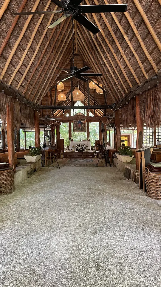Interior de una capilla alta con techo de palma y vigas de madera, piso de grava clara y bancas a los lados, con el altar al fondo iluminado por la luz del jardín