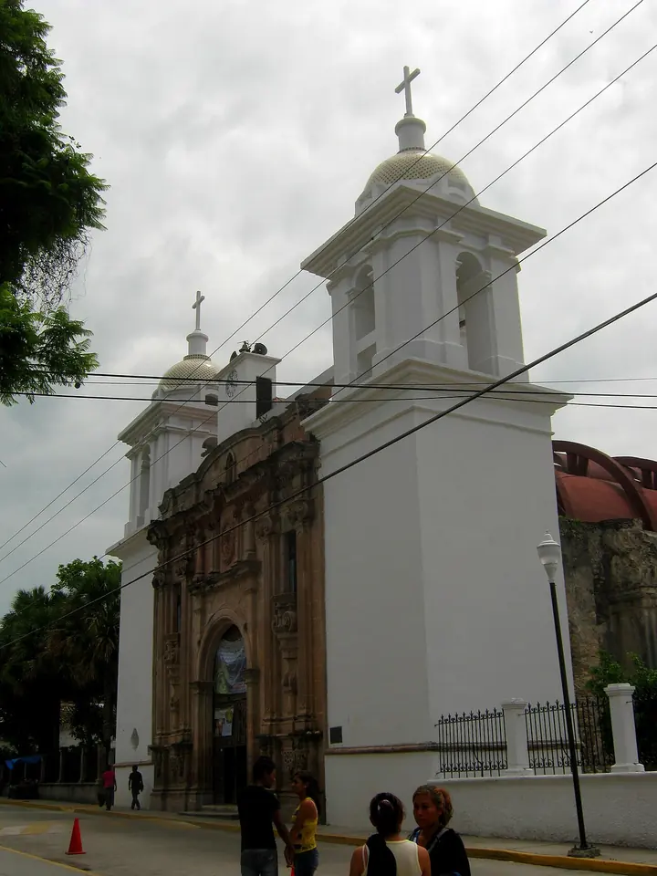 Iglesia colonial blanca con dos campanarios rematados por pequeñas cúpulas y cruces, vista desde la calle con gente que pasa