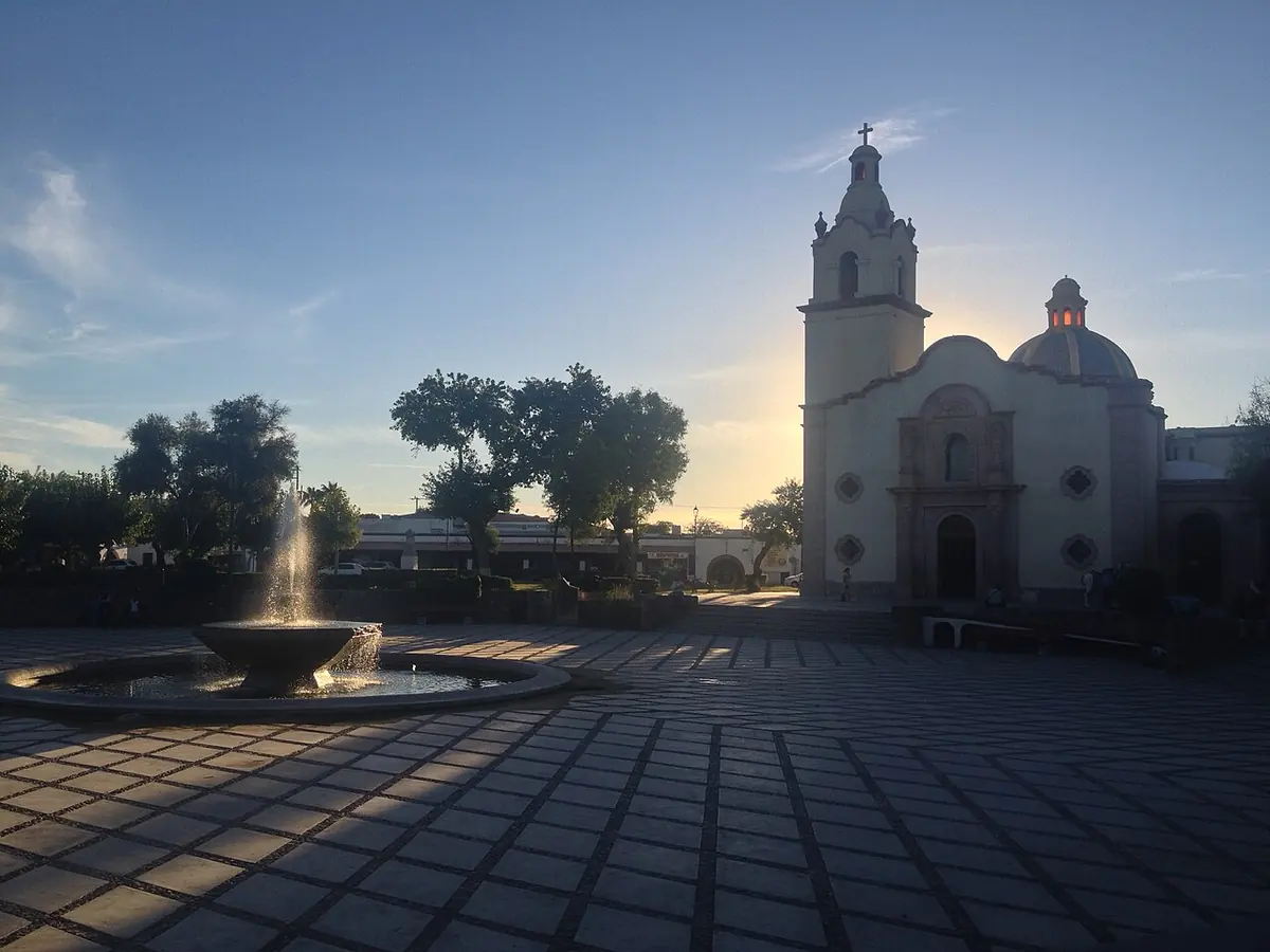 A paved plaza with a fountain at sunset in front of the white church of Santa María Magdalena in Magdalena de Kino