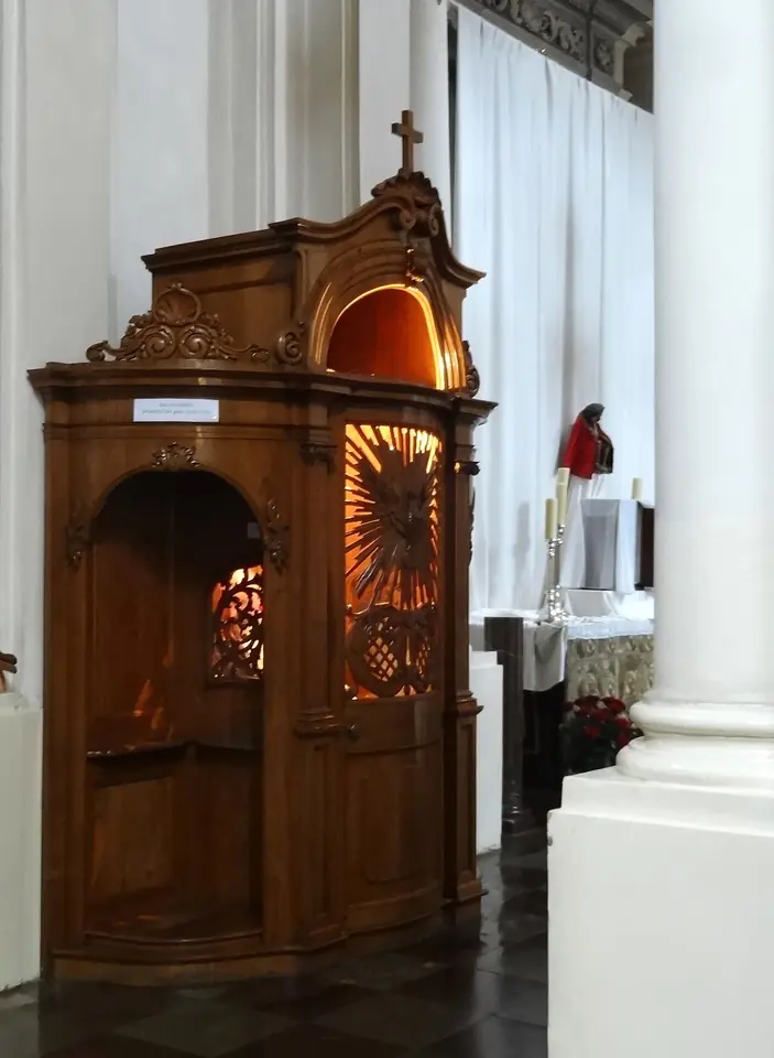 Confesionario de madera labrada, con una cruz en lo alto y una luz cálida encendida dentro, señal de que un sacerdote espera para confesar