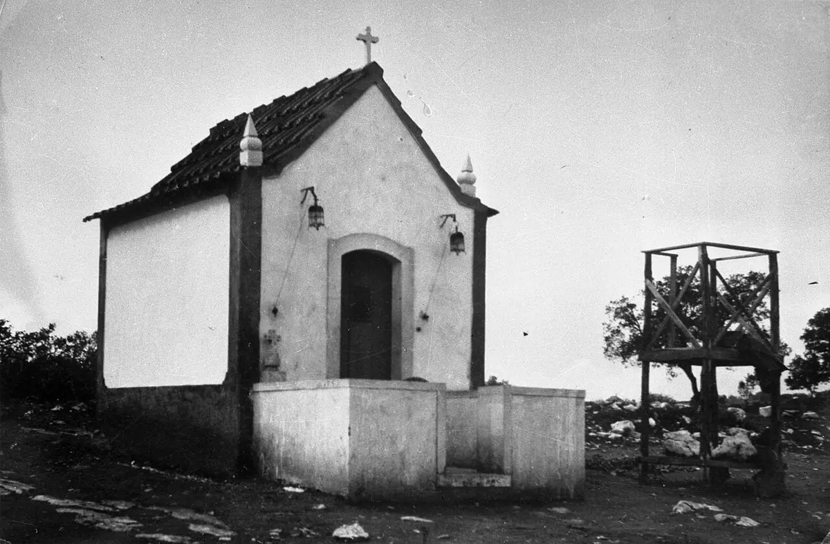 Old black-and-white photograph of a small whitewashed chapel with a tiled roof and a cross, beside a wooden frame on a bare, rocky field