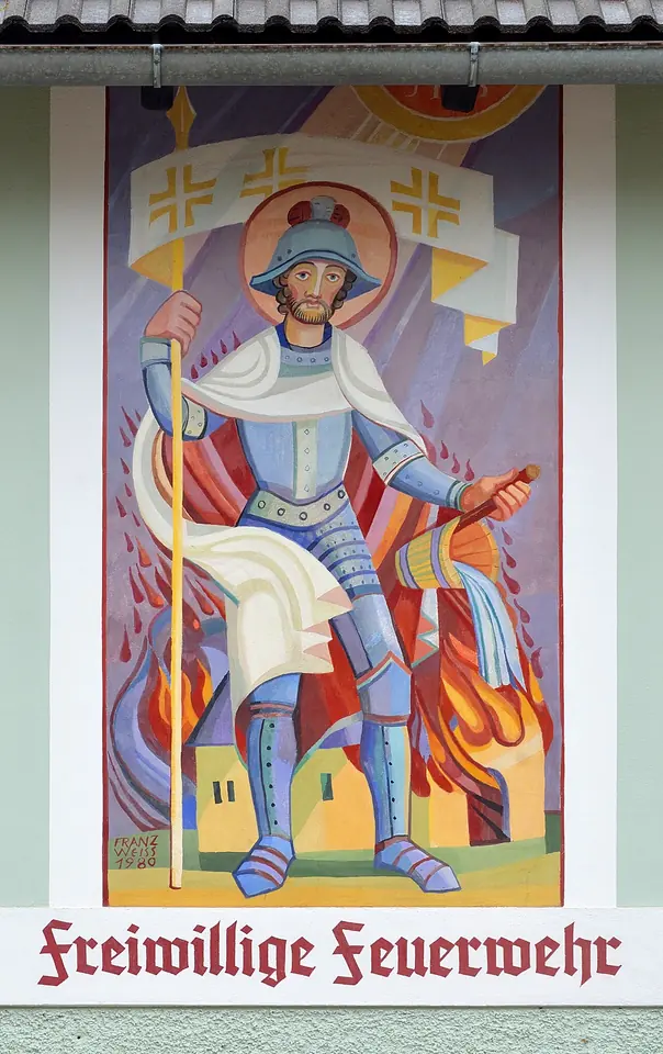 Brightly colored modern painting of St. Florian in armor holding a banner amid flames over a village, above the words Freiwillige Feuerwehr on a fire station wall