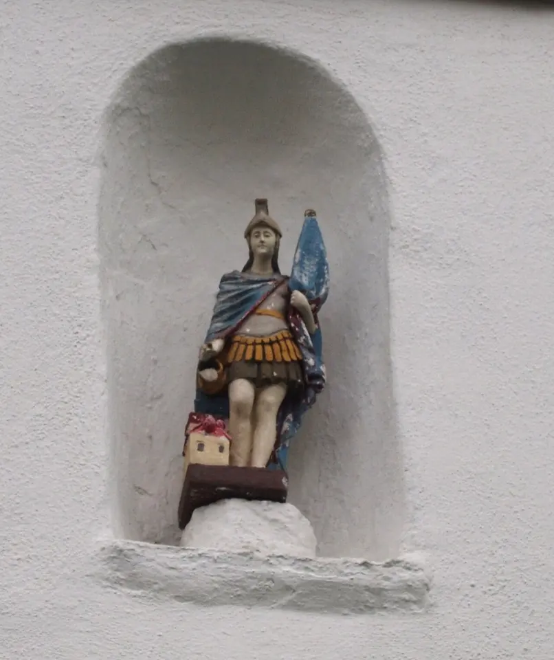 Painted statue of St. Florian in Roman armor and a blue cloak in a white wall niche, holding a banner and pouring water over a small red-roofed house at his feet