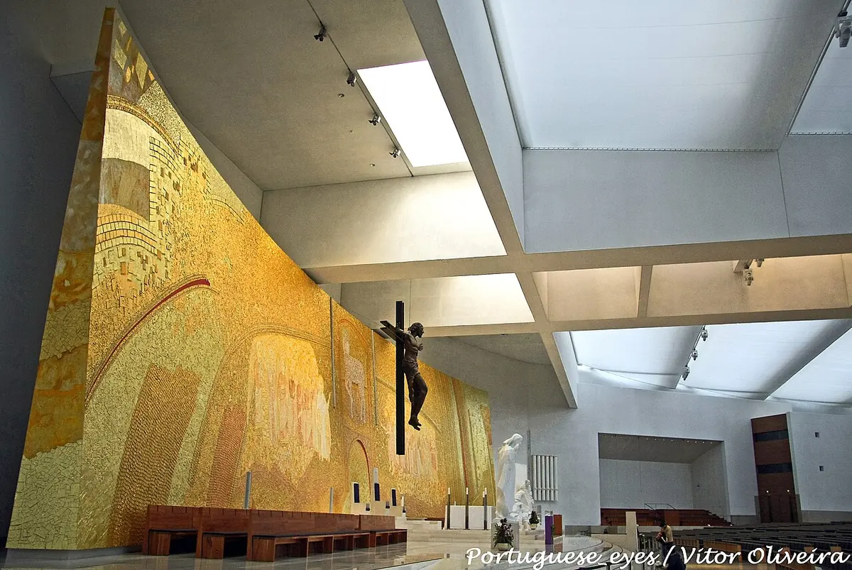 Vast modern church interior with a golden mosaic wall behind the altar, a large crucifix and rows of wooden pews