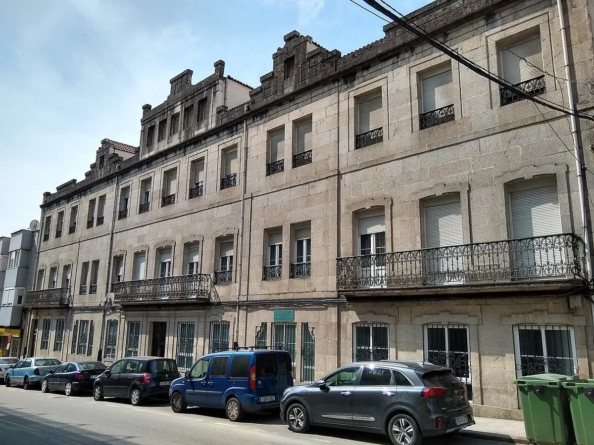 Long three-story stone building with iron balconies along a street in Tui, with cars parked in front
