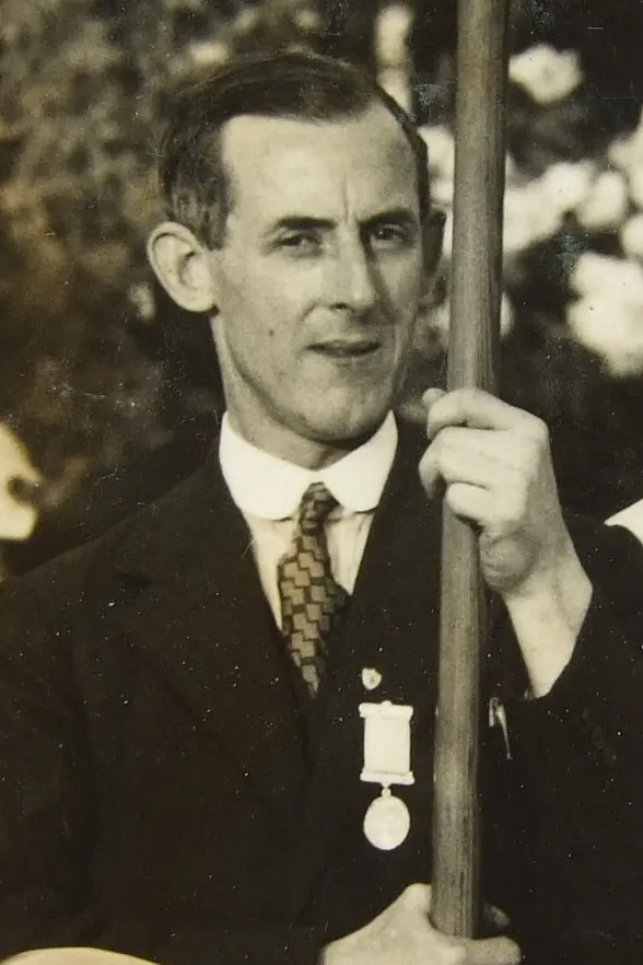 Old sepia photograph of a slim man in a suit and tie with a medal pinned to his chest, holding a banner pole and smiling slightly