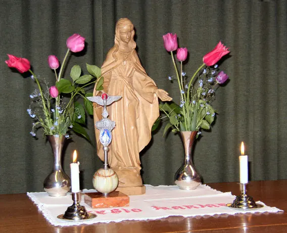 A wooden statue of the Virgin Mary on a white cloth between two lit candles and vases of tulips, with the small standard of the Legion of Mary in front