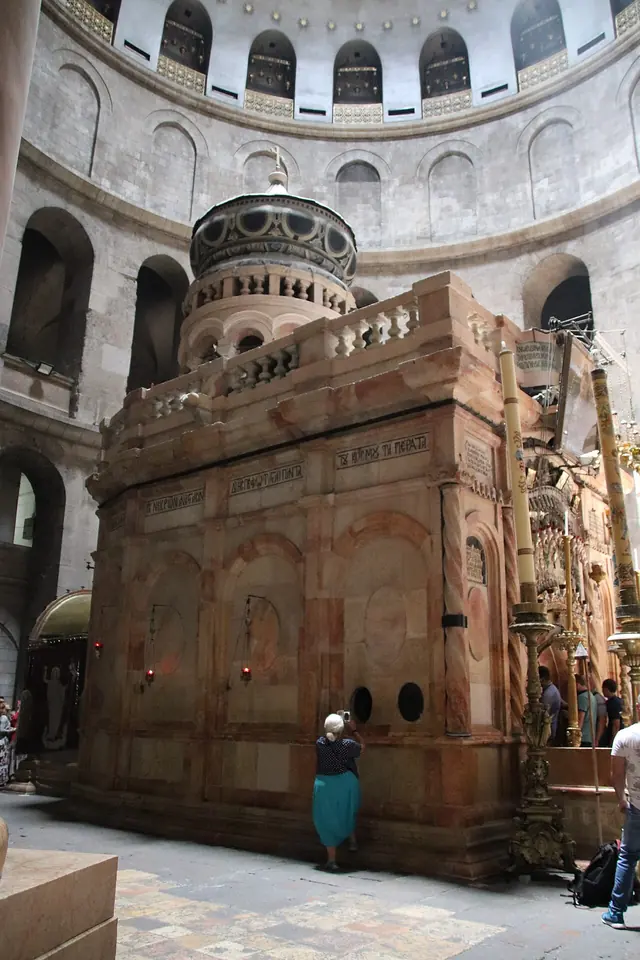 A small ornate stone shrine with columns and hanging lamps stands under a great dome, with pilgrims waiting at its entrance