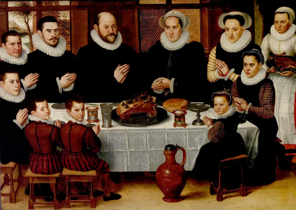 Una familia flamenca numerosa, vestida de negro y con gorgueras blancas, de pie y sentada alrededor de una mesa con un asado y pan, todos con las manos juntas en oración