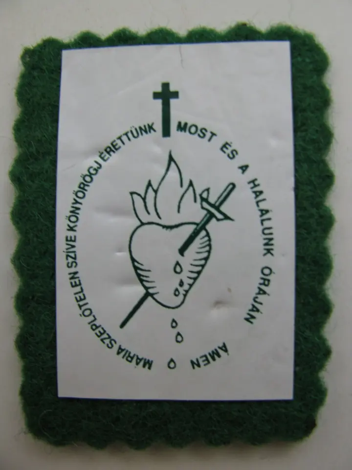 Close-up of a green felt scapular with scalloped edges showing a flaming heart pierced by a sword under a cross, ringed by a Hungarian prayer