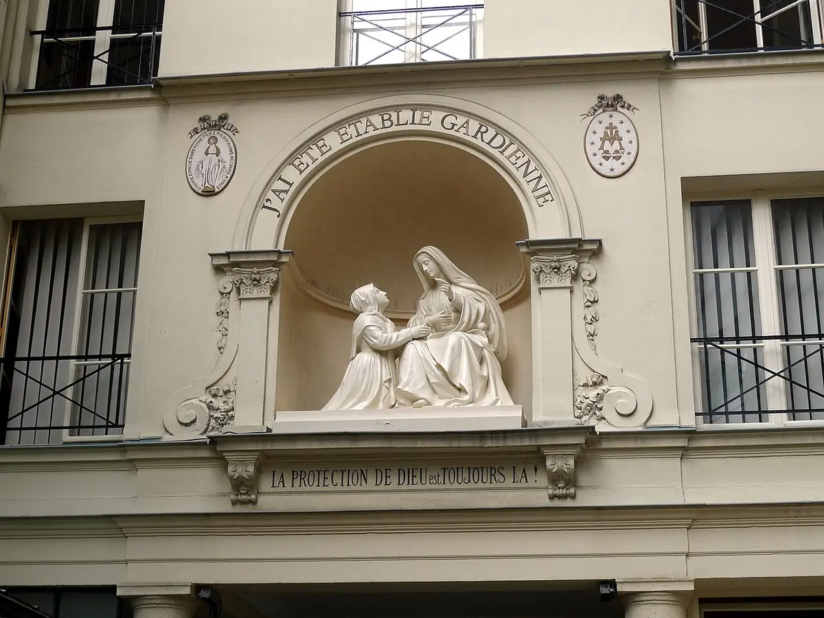 A white stone sculpture in an arched niche above a doorway in Paris, showing a kneeling nun in a winged headdress resting her hands on the lap of the seated Virgin Mary