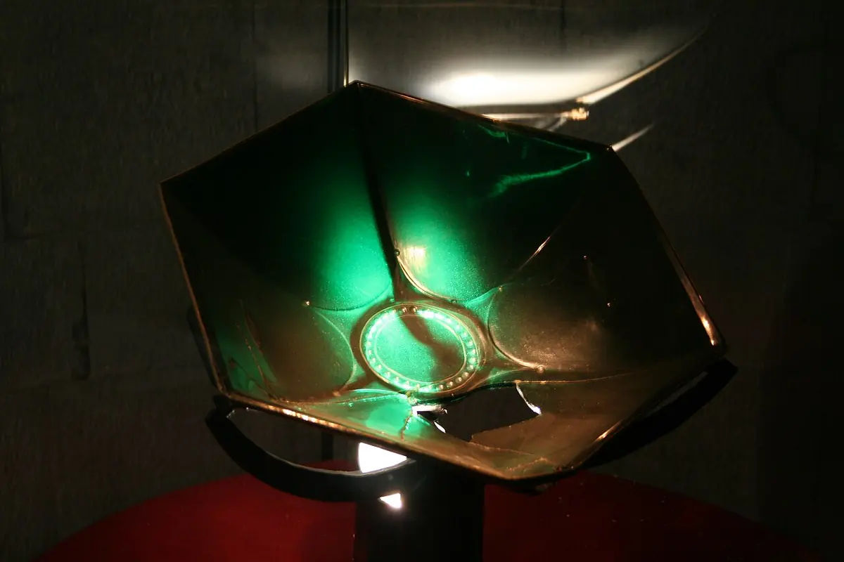 A shallow six-sided dish of green glass, lit from below so that it glows, on a dark stand
