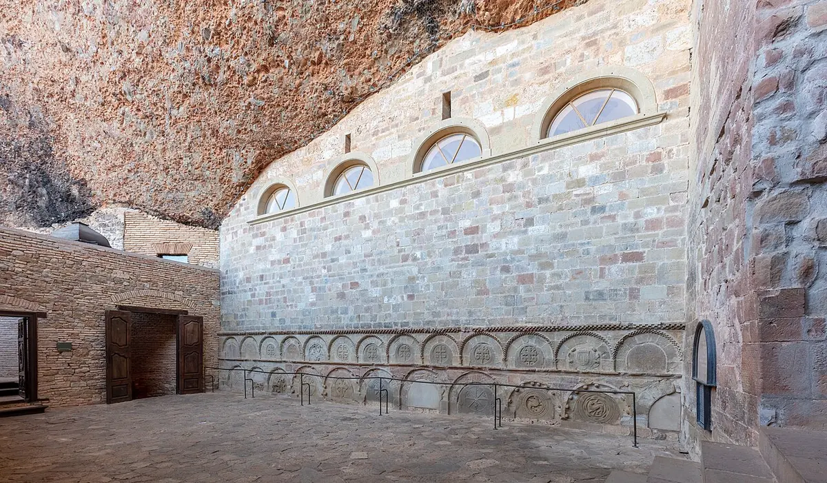 A stone monastery wall with round arched windows tucked under a huge overhanging cliff of reddish rock