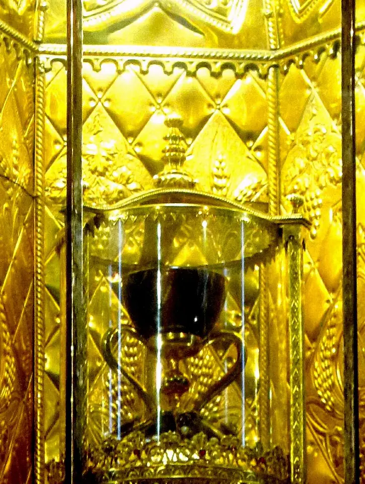 A dark red stone cup on a gold stem with two curved gold handles, inside a glass cylinder against a shining gold background