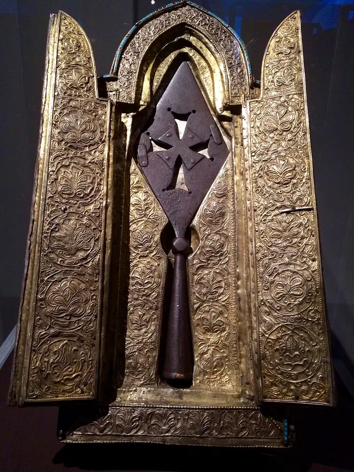 A flat iron spearhead with a small cross cut through it, standing in an open gilded silver case with hinged doors