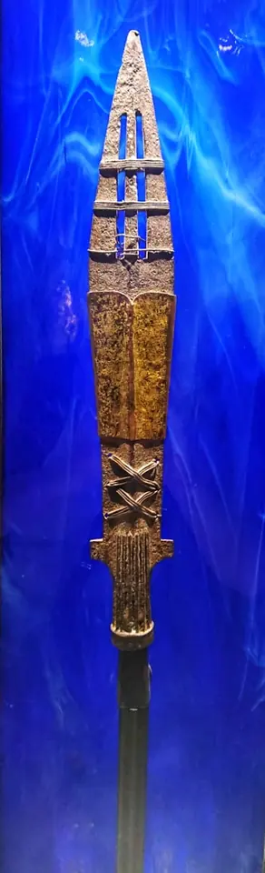 An old iron spearhead with an open slot in its blade and bands of gilded metal, displayed upright against a blue background