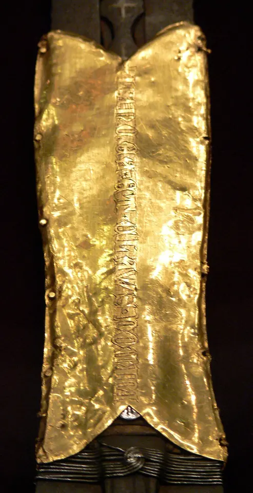 Close-up of a gold sleeve wrapped around the blade of a spear, with a line of engraved Latin capitals running down it
