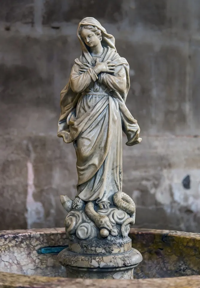 Small marble statue of the Virgin Mary with crossed hands standing on clouds in the middle of a stone holy water basin