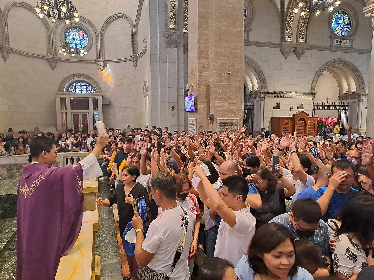 A priest in purple vestments flicks holy water from a sprinkler over a packed crowd of worshipers, many raising their hands, in a large church