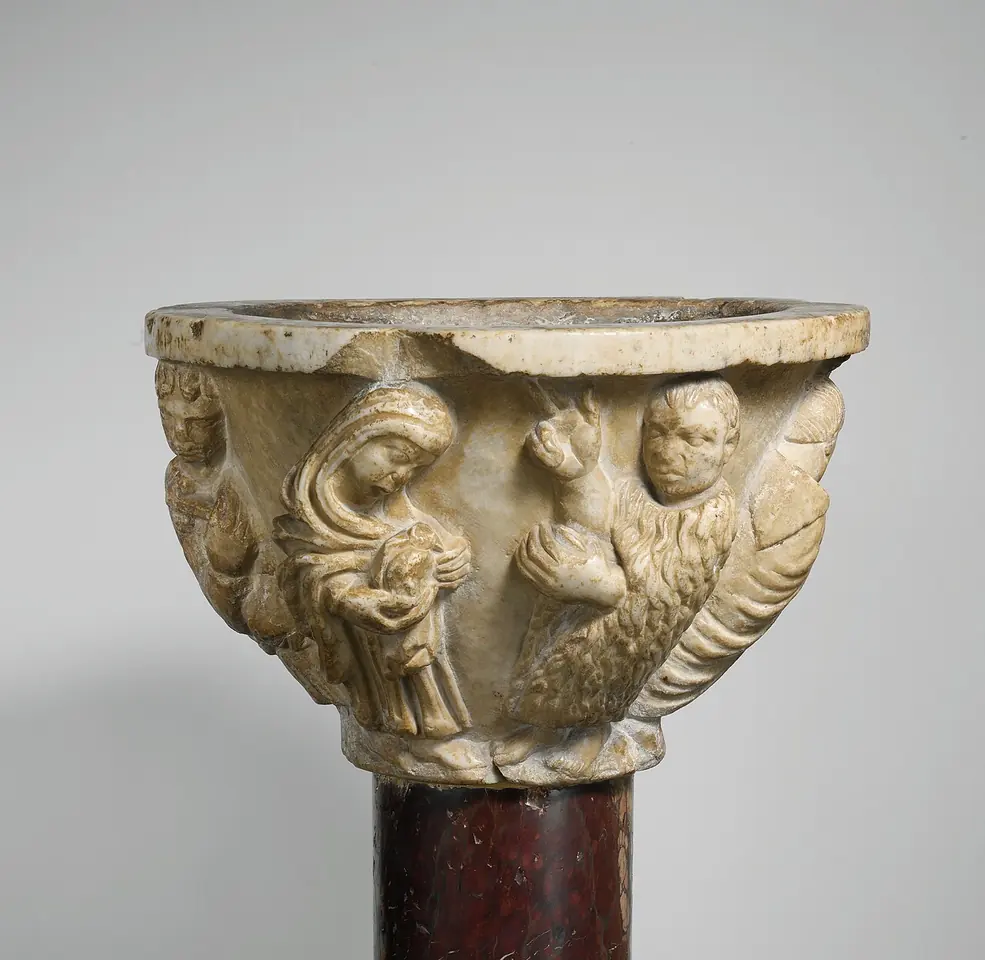 Carved white marble basin on a dark column, with figures in relief around the bowl, including a veiled woman and a winged creature