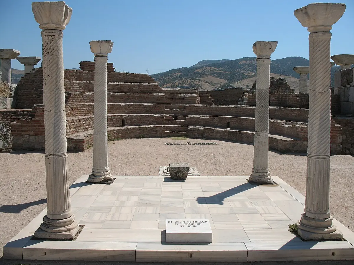 A square marble platform marked by four tall columns in the open air, with a small plaque, among the ruins of an ancient basilica