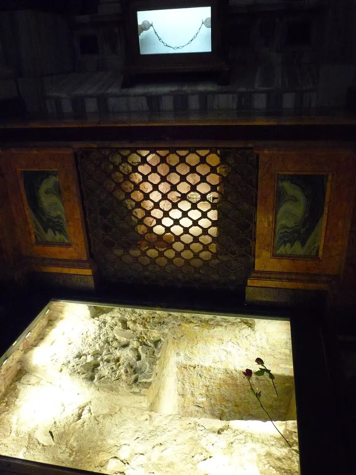 A lit opening in the floor showing ancient stonework below a grille, with a chain displayed in a glass case above