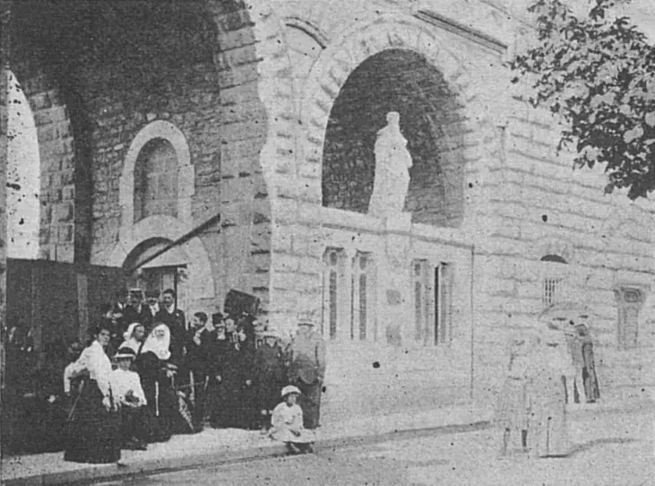 Old photograph of the low stone building of the Lourdes Medical Bureau under a rampart arch, with nuns, women and children gathered at its door