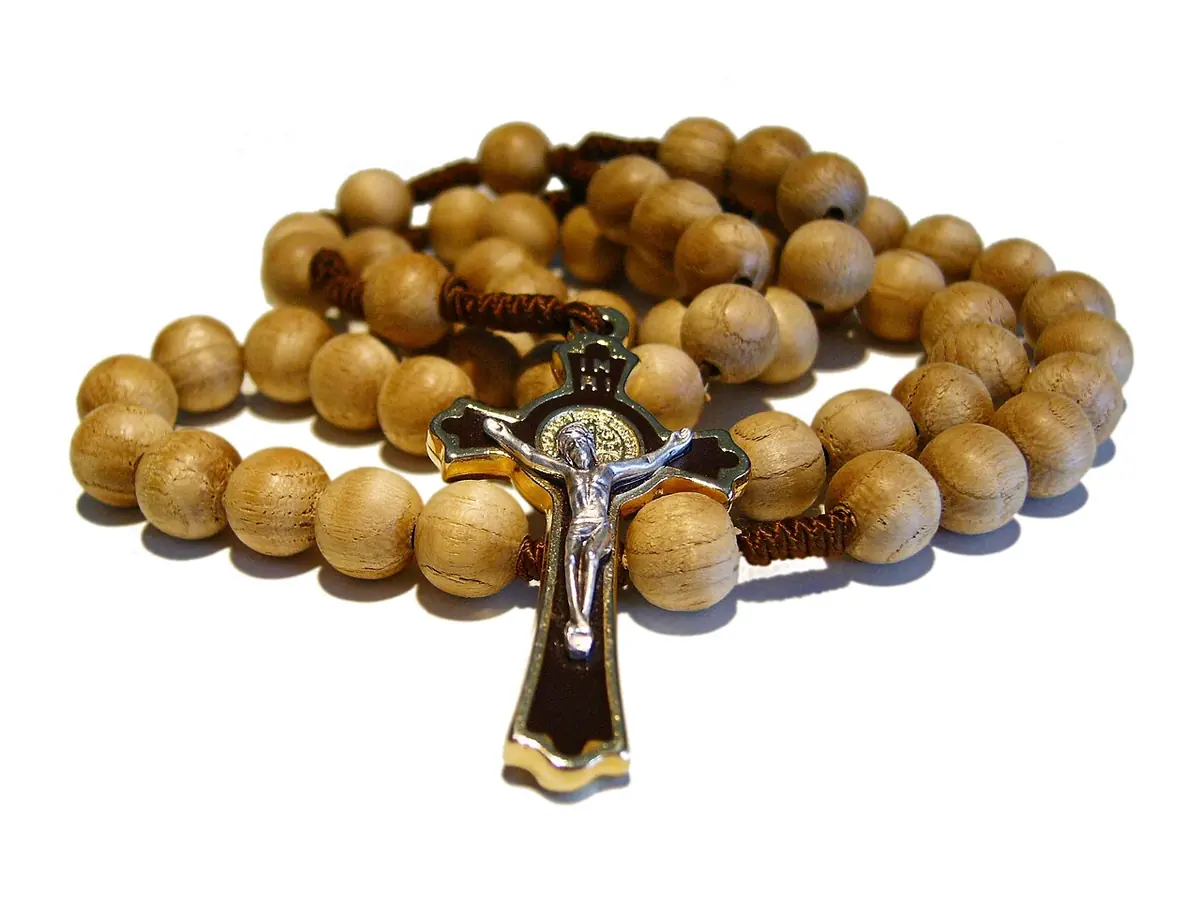 A rosary of round light-wooden beads on a brown cord, coiled on a white surface, with a dark metal crucifix in front