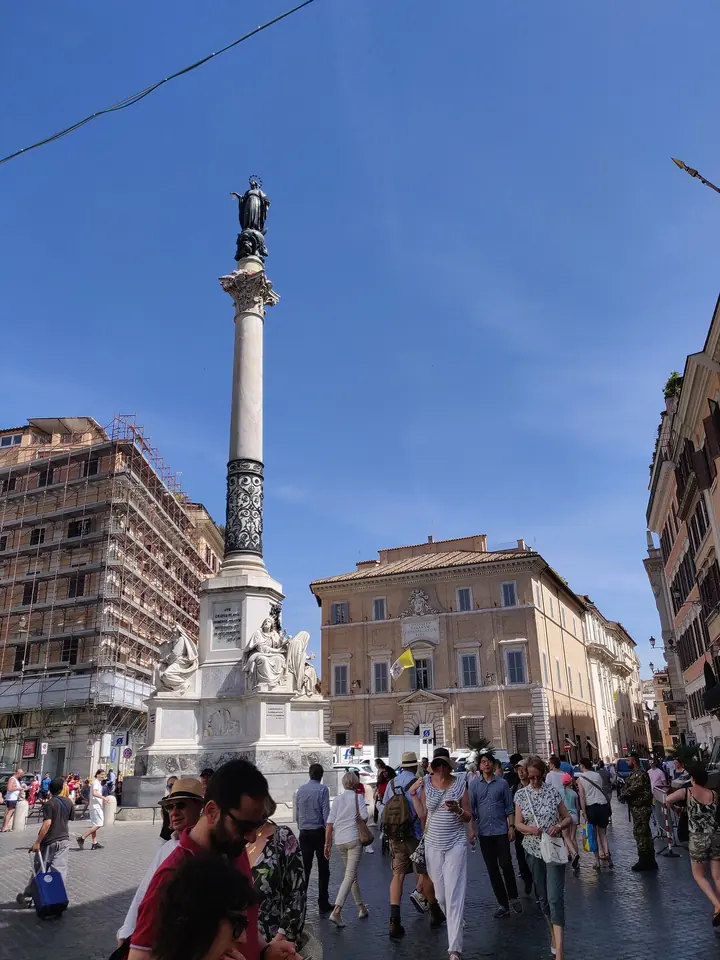 A tall marble column topped by a bronze statue of Mary against a blue sky, rising over a busy Roman square with palazzi on both sides