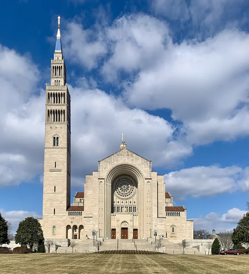 The pale stone front of the National Shrine in Washington, with a great round arch, a rose window, a dome and a tall bell tower under a blue sky with white clouds