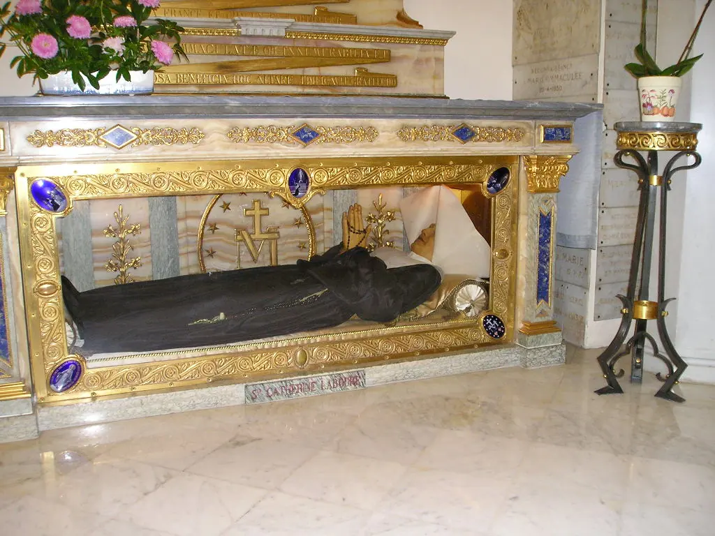 A nun in a dark habit and white collar lying in a glass and gilded reliquary under a marble altar, with flowers above