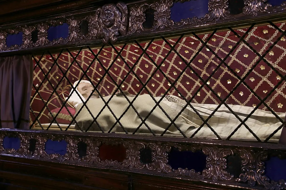 A figure in a pale habit lying behind a silver lattice grille in an ornate shrine of silver and red