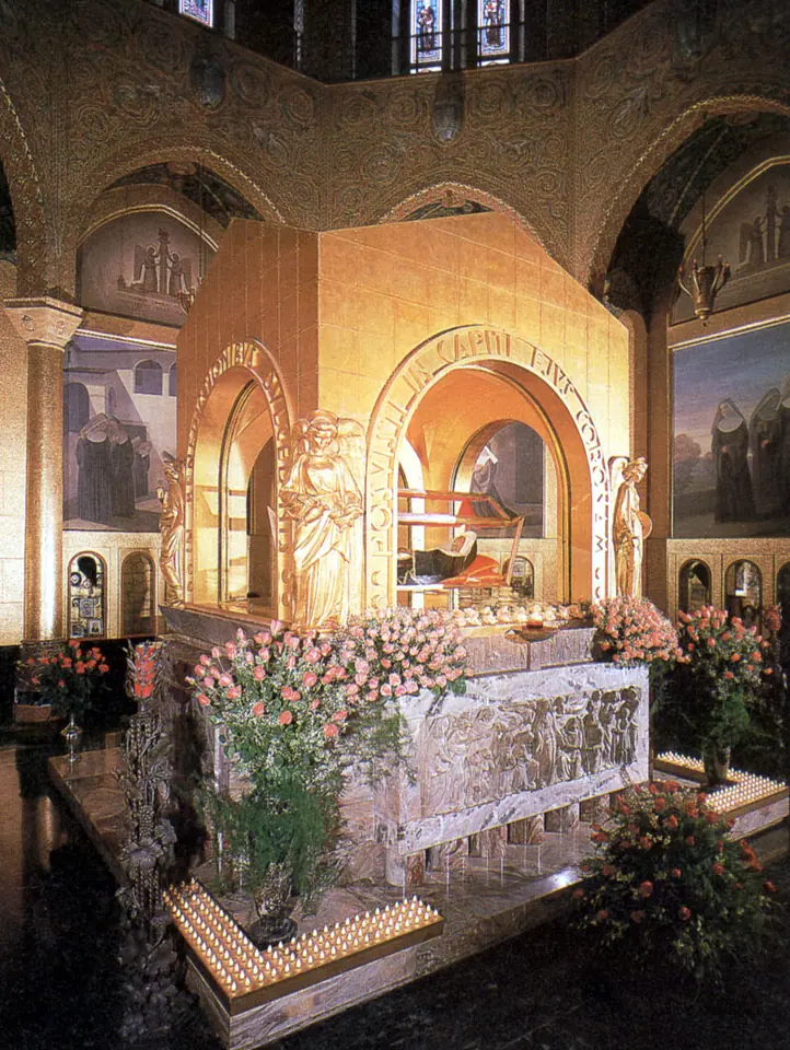 A gilded shrine with glass panels in a chapel decorated with paintings, surrounded by pink roses and rows of candles