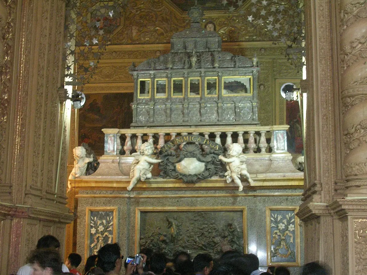A tall silver casket with small windows on a carved marble and jasper base, decorated with cherubs, in a gilded chapel with visitors in front