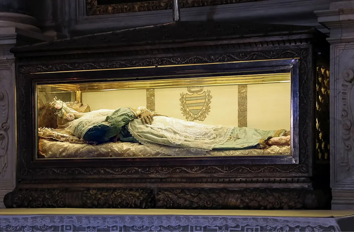 A small darkened body in a light gown and veil lying in a glass case set into a dark carved wooden altar