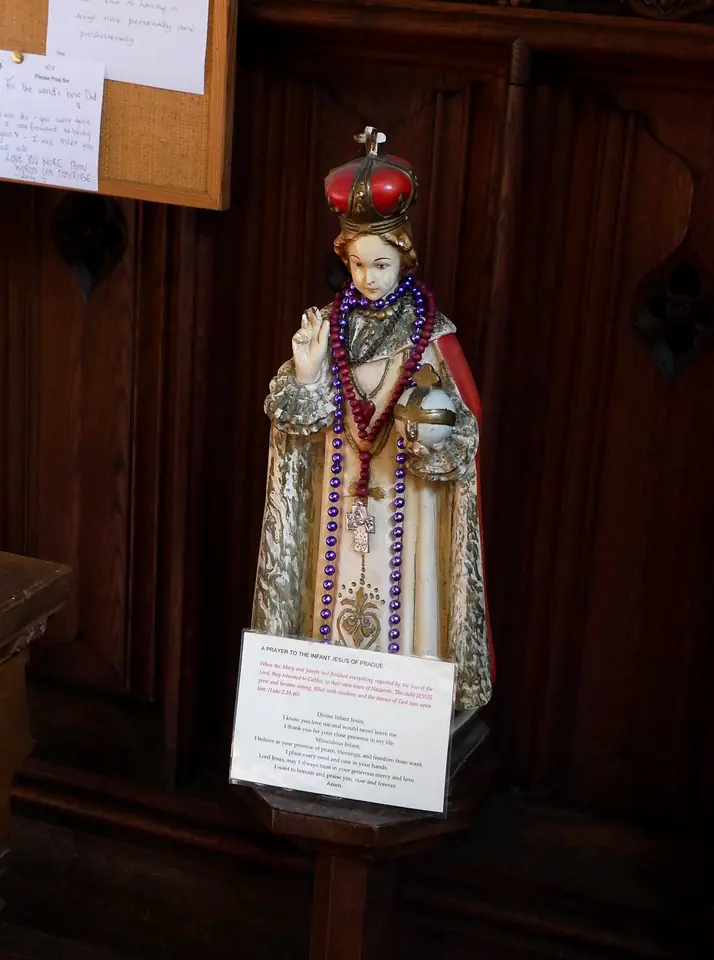 Infant of Prague statue in a white and silver robe and red crown, hung with rosaries, standing against dark wood paneling beside a handwritten prayer card