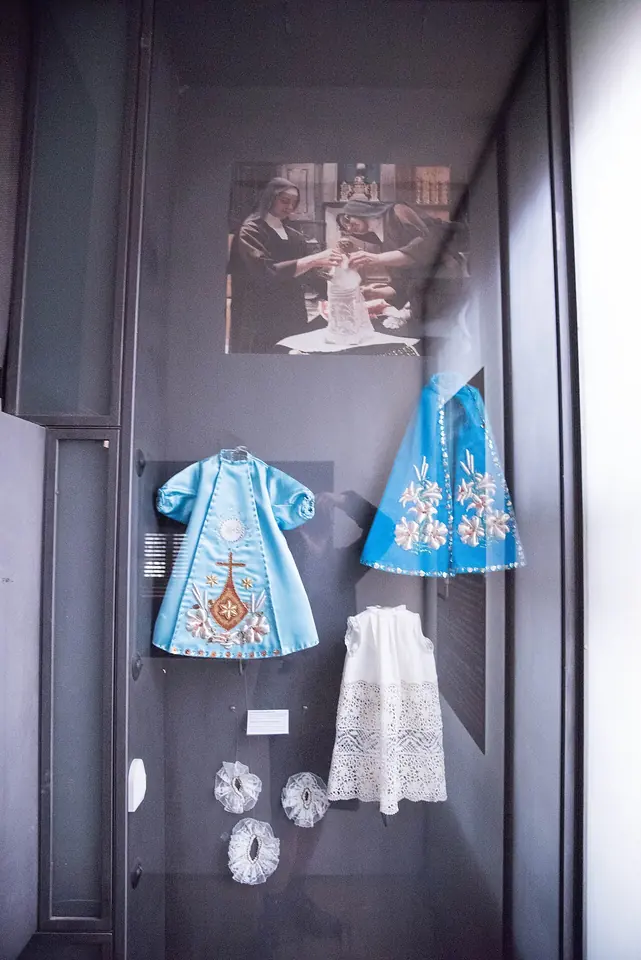 Glass case holding two small light-blue embroidered robes, a white lace alb and lace collars made for the Infant of Prague statue, below a photograph of two nuns dressing the statue