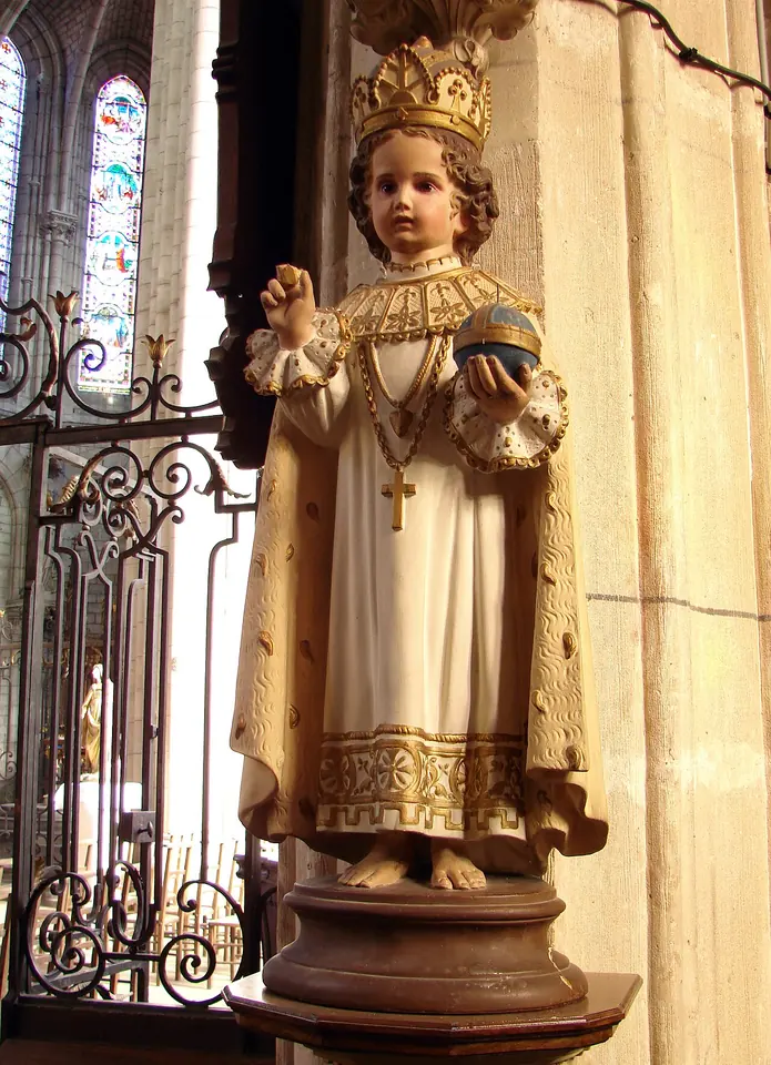 Painted statue of the Infant of Prague in a cream robe and ermine-trimmed mantle with a gold crown, raising his hand in blessing and holding a blue globe, beside an iron grille in a church