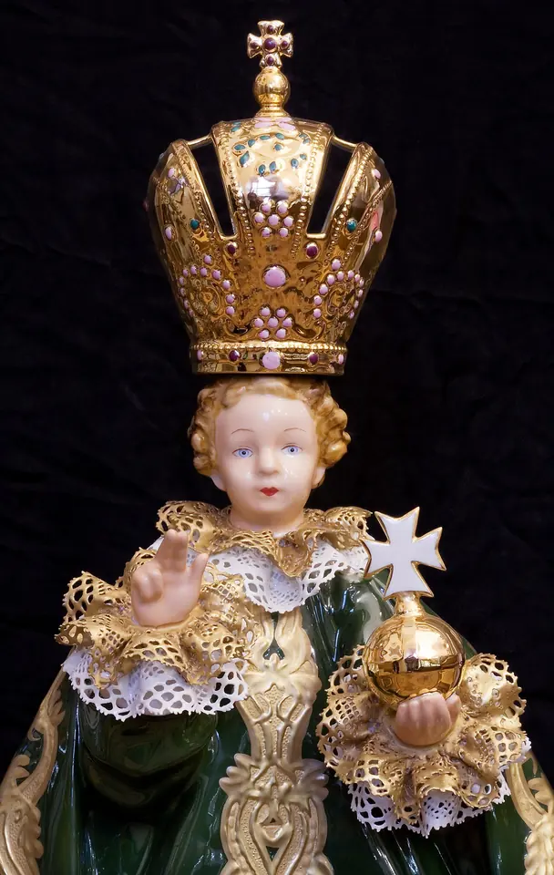 Close view of an Infant of Prague statue in a green robe with gold lace, wearing a tall jeweled crown and holding a gold orb topped with a cross
