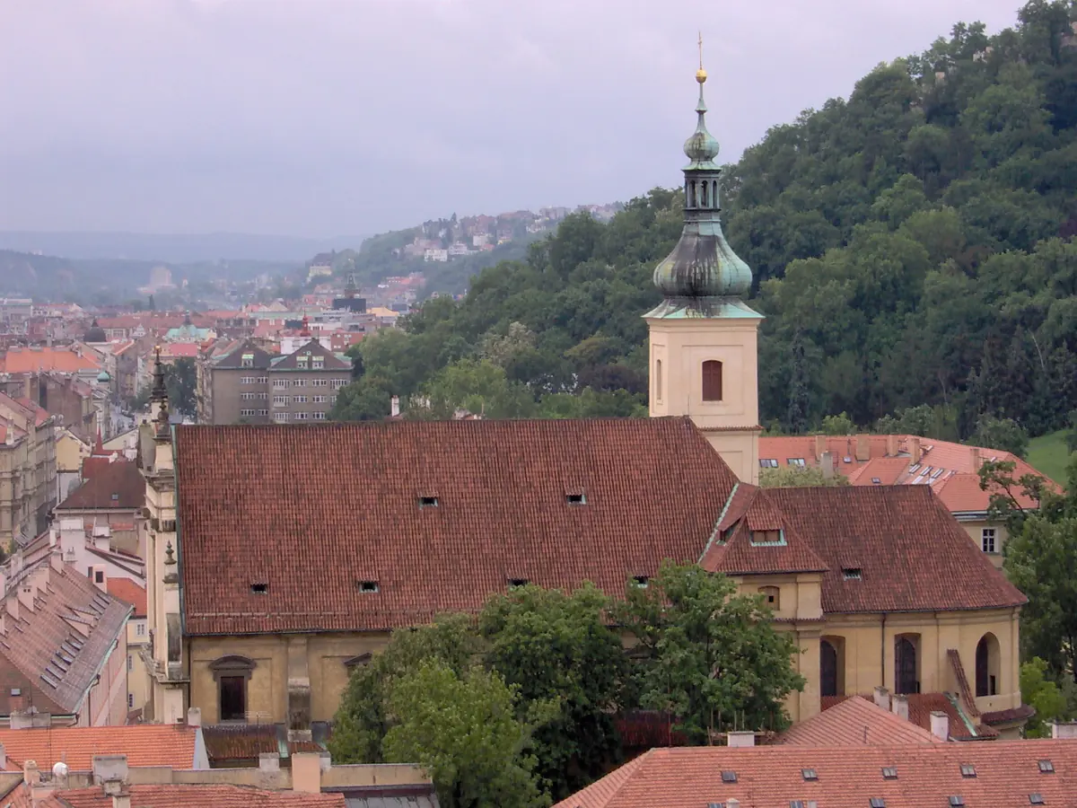 A view over red tile roofs in Prague's Lesser Town to the long roof and single bell tower of the Church of Our Lady Victorious, with the green slope of Petřín Hill behind