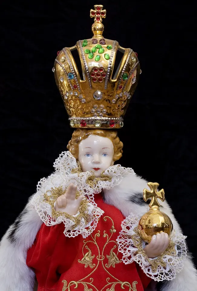 A painted statue of a curly-haired child in a plain white tunic, raising two fingers of his right hand in blessing and holding a blue and gold orb with a cross in his left
