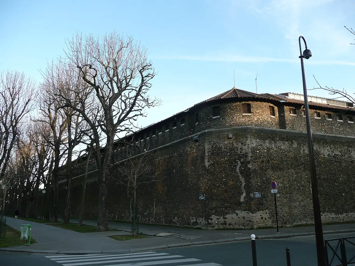The high, rough stone outer wall of La Santé prison curving along a Paris boulevard lined with bare trees, under a blue winter sky
