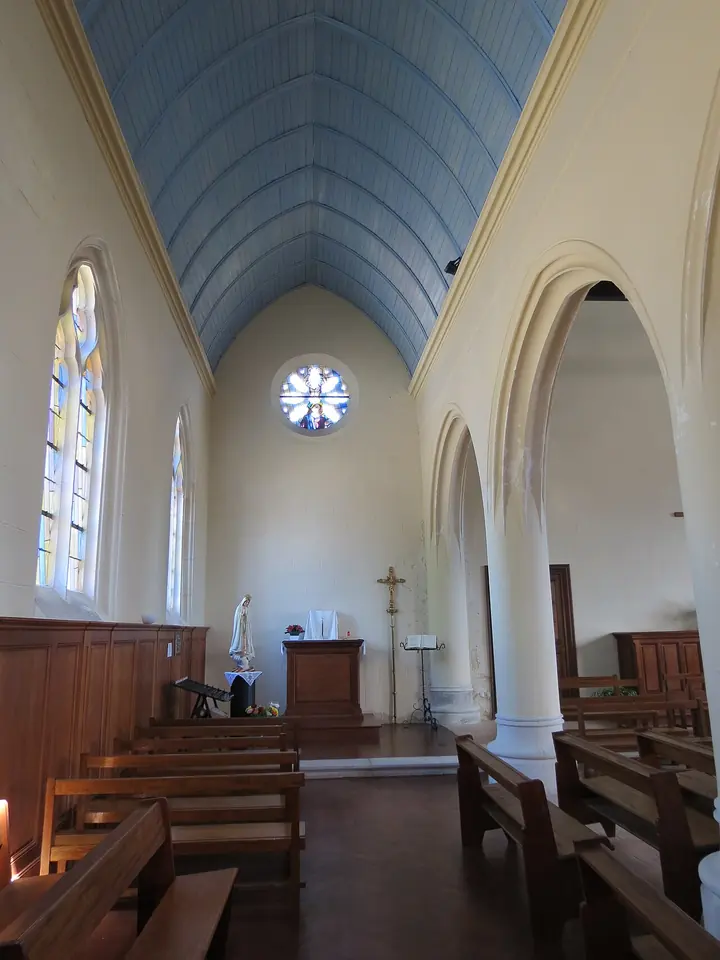 Simple church interior with pale walls, a blue-painted wooden vault, round arches and wooden pews leading to a plain sanctuary under a rose window