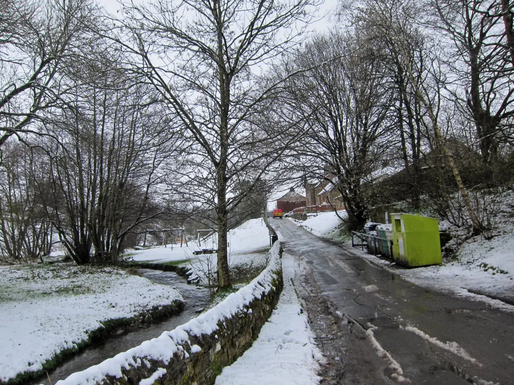 A narrow village lane in the snow beside a stream lined with bare trees, leading to a few houses in the distance