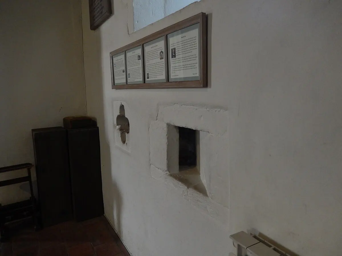 Inside wall of an old stone church with a small four-lobed opening and a narrow slanted slit, through which an enclosed anchoress could see the altar
