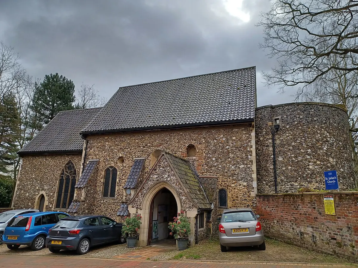 Small flint church with a tiled roof and a round tower, with a modern chapel built onto its side, seen from a parking area under a cloudy sky