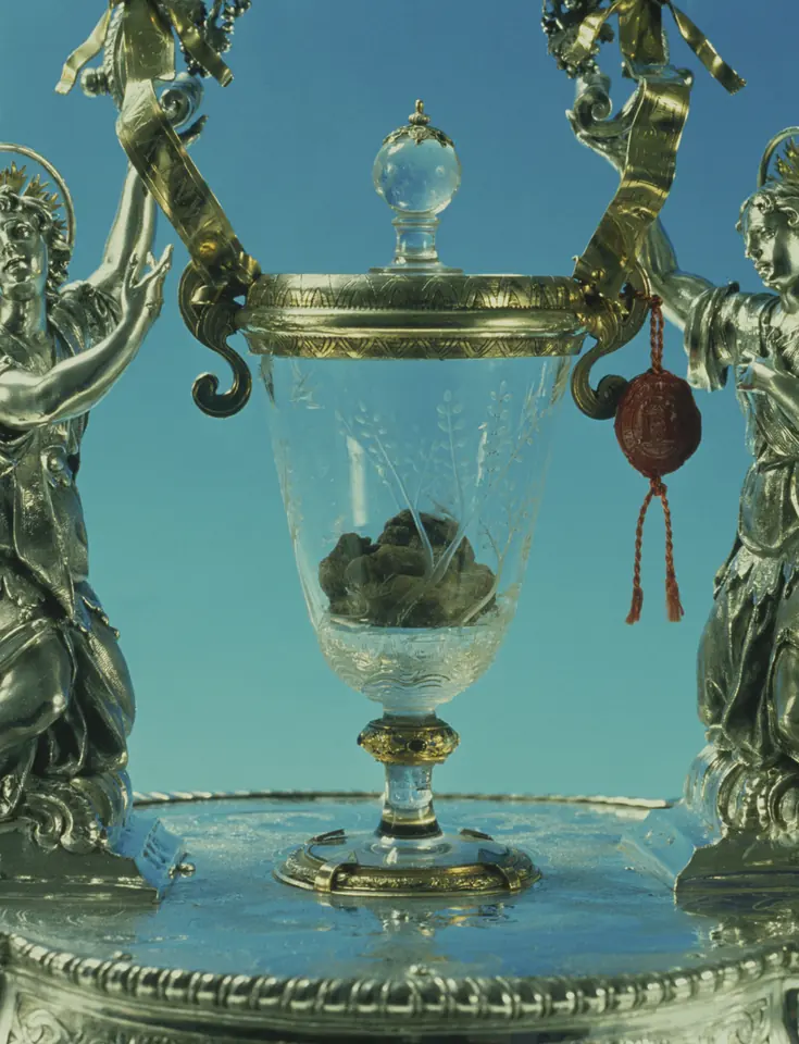 A close view of a clear crystal chalice with a lid, holding small dark brown lumps, held up by two silver angel figures, with a red wax seal on a cord